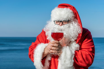 Fat man in glasses dressed as Santa drinking wine on the ocean. Funny and happy