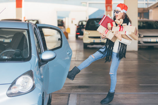 Young woman opening door of her car with leg (foot) while her hands are full of christmas gifts and presents boxes, after xmas shopping in mall