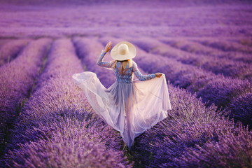 Young woman runs in purple lavender field sunset. Dress and hat develop wind. France, Provence.