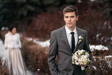 Handsome young man with bouquet in hands in winter park. His bride is standing on a blurred...