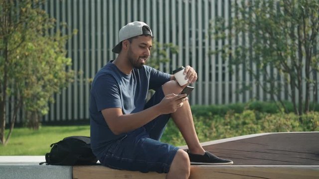 Smiling young man wearing baseball cap and jeans shorts is drinking coffee and web surfing on his phone sitting on bench in the park. Left to right pan slow motion medium shot