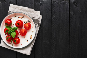 Top view of tomatoes in a plate with Himalayan salt, pepper and basil on gray dishtowel over black wooden table. Top view with copy space for menu or recipes. Food background concept.