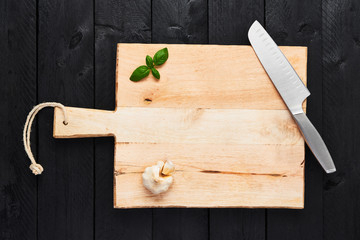 Top view of chopping board with stainless steel vegetable knife, garlic and basil on black wooden background with copy space.