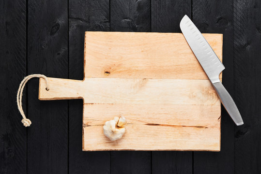 Top View Of Chopping Board With Stainless Steel Vegetable Knife And Garlic On Black Wooden Background With Copy Space.