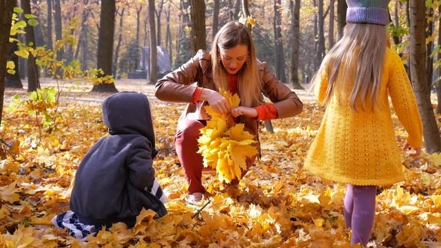 Mother knits a yellow leaf wreath together with two children at park in autumn with colorful leaves and trees background