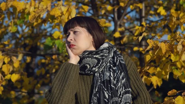 A young sick woman in a green knitted sweater strokes her head against the background of yellow foliage in the city park in Indian summer.