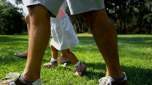 Young parents with child walking on sunny lawn, cropped view. Mother, father and daughter spending time together in park. Family walk concept