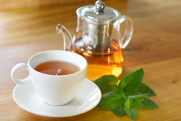White cup and glass teapot of herbal tea with fresh mint twig on wooden table