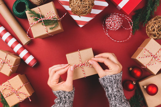 Christmas background with gift boxes, clews of rope, paper's rools and decorations on red. Preparation for holidays. Top view with copy space. Woman's hands holding gift box.