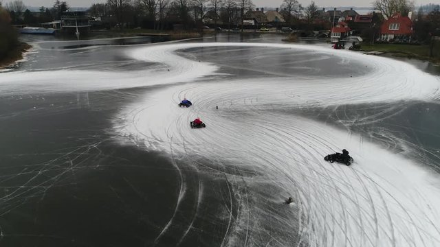 Aerial footage of go-karts drifting through corners of frozen lake ice track showing the two karts in competition trying to stay ahead then driving away behind tree line 4k high resolution quality