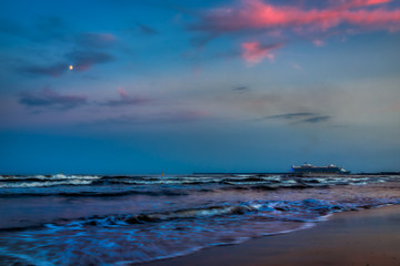 Waves crashing on the shore with a dark blue sky and the bright pinks of the sunset with a cruise ship on the horizon and the hazy moon in the distance