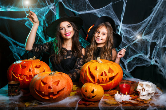 Happy Halloween . Beautiful caucasian mother and her daughter in witch costumes enjoy using magic with magic wand to halloween pumpkin jar over spider web on black studio background.