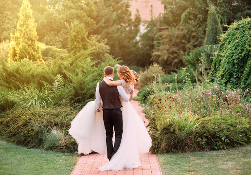 young groom in a suit carries in arms his bride, wearing a long magnificent luxurious white dress, walking at amazing garden. no faces. picture taken from the back. beautiful romantic photo, art