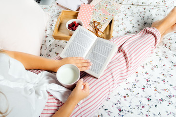 Young woman drinking milk at home in bed and reading book, top view