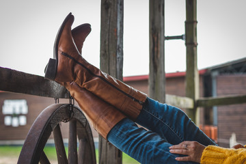 Woman is relaxing on the ranch with her legs on a wooden railing. Leather boot and jeans