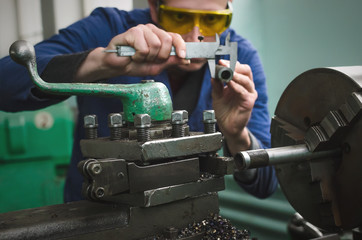 Turner is working on a lathe machine in a factory. Worker is measuring a produced detail by caliper.
