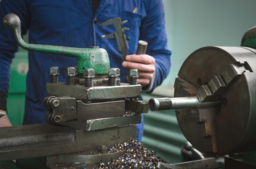 Turner is working on a lathe machine in a factory. Worker is measuring a produced detail by caliper.