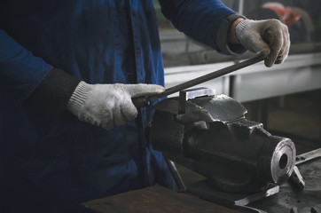 Man worker is working with iron detail on vise grip on blacksmith table and using a file.