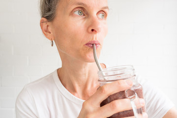 Closeup of middle aged woman drinking berry smoothie from glass mug with metal straw against white background (selective focus)