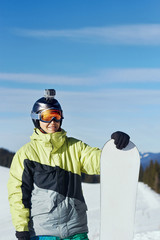 Fototapeta na wymiar .Snowboarder with action camera on a helmet. Ski goggles with the reflection of snowed mountains. Portrait of man at the ski resort on the background of mountains and blue sky, hold snowboard.