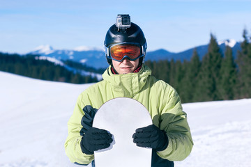 Fototapeta na wymiar .Snowboarder with action camera on a helmet. Ski goggles with the reflection of snowed mountains. Portrait of man at the ski resort on the background of mountains and blue sky, hold snowboard. 