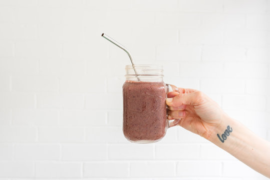 Woman's hand with love tattoo on wrist holding berry smoothie in glass mug with metal straw against white background  (selective focus)