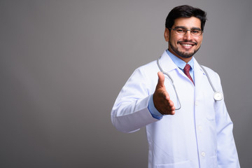 Young handsome bearded Persian man doctor against gray backgroun