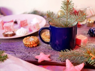  On a wooden table, a branch of spruce in a blue cup on the background of a pink dish with sweets, seasonal winter home holidays, Christmas, New Year