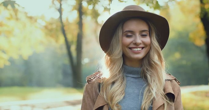 Close up of the charming young blond Caucasian woman in the hat smiling to the camera in the middle of the picturesque park in the fall season. Portrait.