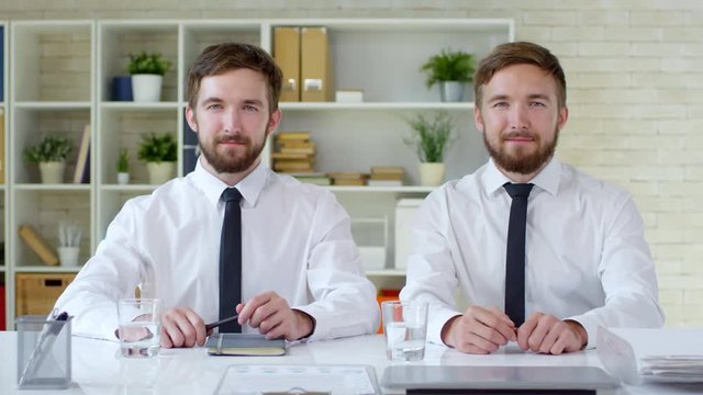 Portrait of two optimistic bearded Caucasian twin brothers in formal clothes sitting next to each other at desk in office and smiling at camera, medium shot