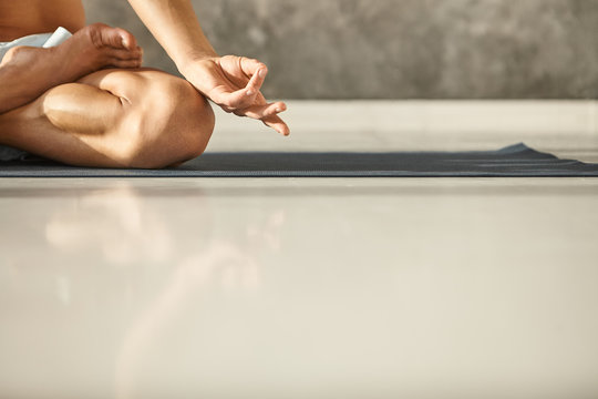 Horizontal cropped shot of unknown male with tanned athletic body, practicing meditation on yoga mat with legs crossed in lotus pose. Unrecognizable guy meditating at gym. Close up on man's hand