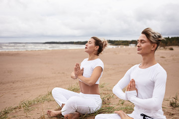 Two atheltic European people man and woman in white sportswear closing eyes and holding hands in namste gesture, sitting in padmasana while meditating outdoors during yoga retreat by the ocean