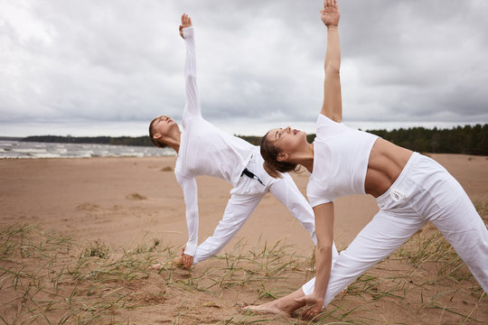 Outdoor portrait of attractive woman and young male with athletic bodies both dressed in white outfits, practicing yoga at seaside during retreat, doing Utthita Trikonasana or Extended Triangle pose