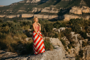 A beautiful blonde girl in the red dress posing in the ancient canyon on the sunset. Spain