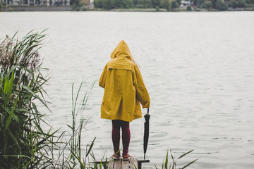 cold fresh morning foggy weather, autumn season, person back to camera in yellow rain coat and black umbrella in right hand on small wooden pier near river