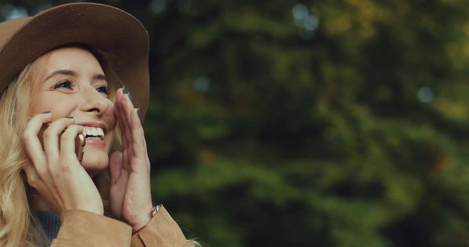 Close up of the happy cute young Caucasian woman in a hat having a mobile phone talk and laughing laud and cheerfully. Outdoor.