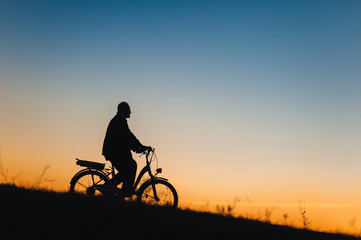 Fototapeta na wymiar Male cyclist on the e-bike or electric bicycle on the sunset background slides down the hill. Silhouette of the old man in profile. Active pension. Travel. Sport.