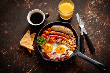 English breakfast in iron cooking pan with fried eggs, sausages, bacon, beaked beans, toasts, orange juice and black coffee on dark rusty background.