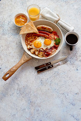 Full English Breakfast served in a pan. Fried eggs, beked beans, tomatoes, champignons, crispy bacon, sausages and toast. Placed on stone background. Top view with copy space.
