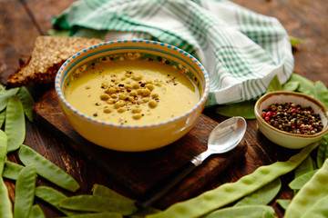 Green vegetables cream soup in grey bowl placed among fresh peas and bean pods with full grain bread on wooden background. Top view with copy space.