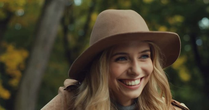 Portrait of the cheerful blonde stylish girl in a hat laughing in front of the camera in the park in autumn. Close up.