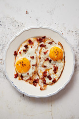Two fresh fried eggs with crunchy crisp bacon and chive served on white ceramic plate. Fork and knife on side. Top view.
