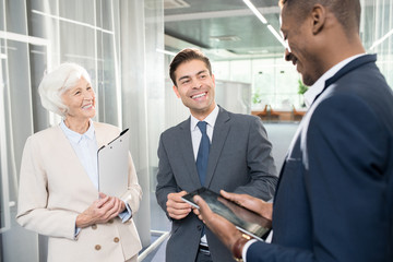 Cheerful optimistic business partners in formalwear standing in modern office and laughing while talking about project, African-American businessman holding tablet and sharing ideas with colleagues