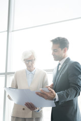 Serious confident senior lady boss in glasses reading papers in folder and commenting report data while talking to employee in office corridor