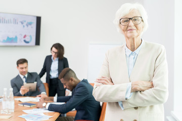 Content confident attractive senior chief executive officer in glasses wearing jacket standing in meeting room and looking at camera while business analysts working with data in background.