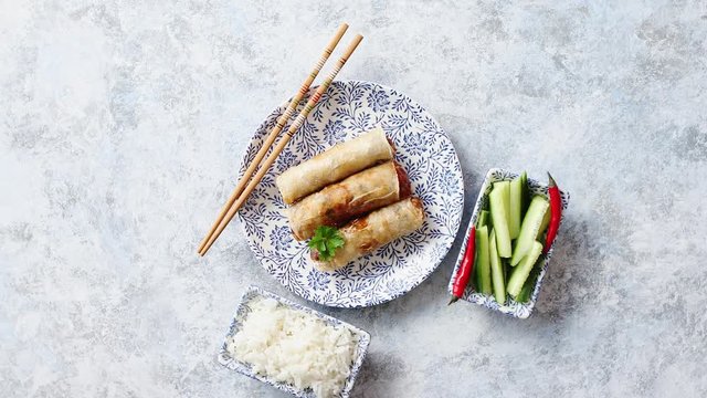Orginal asian deep fried spring rolls placed on beautiful oriental style plate with chopstick, boiled rice, cucomber nad hot chilli peppers od side. Top view on stone background.