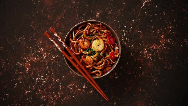 Traditional asian udon stir-fry noodles with shrimp in bowl and chopsticks. Fresh chilli pepers on side. Placed on dark rusty background with copy space.