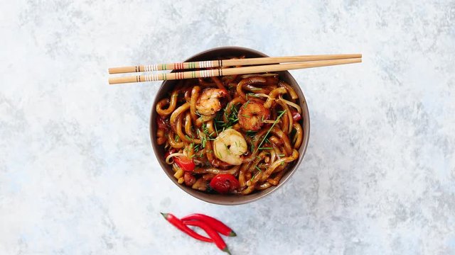 Traditional asian udon stir-fry noodles with shrimp in bowl and chopsticks. Fresh chilli pepers on side. Placed on bright stone background with copy space.