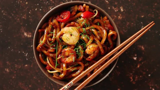 Traditional asian udon stir-fry noodles with shrimp in bowl and chopsticks. Fresh chilli pepers on side. Placed on dark rusty background with copy space.