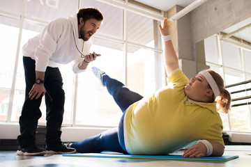 Full length portrait of obese young woman stretching legs while working out with personal coach in fitness club